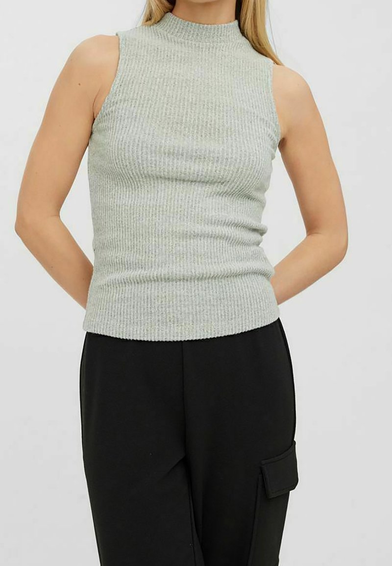 Femme portant un haut en maille côtelée sans manches gris clair et un pantalon noir taille haute avec une poche latérale, debout devant un fond uni.