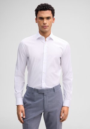 Mann mit lockigem Haar, der ein eng anliegendes weißes Hemd und hellgraue, maßgeschneiderte Hosen trägt und vor einem schlichten Hintergrund steht.