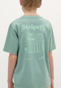 T-shirt en coton vert avec des manches courtes, présentant des graphiques imprimés en vert clair d'une façade de cinéma et du texte, incluant "Scalpers. Films."