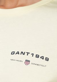 Hellgelbes Baumwoll-T-Shirt mit Rundhalsausschnitt, bedruckt mit dem Schriftzug "GANT1949" in kräftigem Schwarz und einem rot-weiß-blauen Schild-Logo.