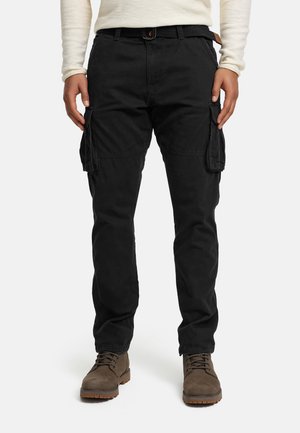Homme portant un pantalon cargo noir avec des poches à l'avant et sur les côtés, une chemise beige à manches longues, et des bottes marron à lacets, debout face à l'avant.