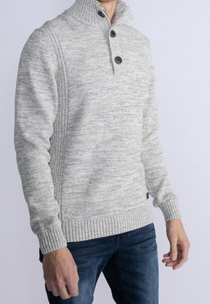 Grå striktrøje med høj krave, tre sorte knapper og ribbede manchetter. Struktureret mønster gennem hele stoffet. Båret med blå jeans.