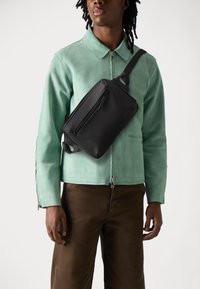 Sac à bandoulière noir en matériau lisse, avec une poche zippée à l'avant et une bandoulière réglable, porté sur une veste en daim verte.