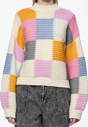 Pull en maille épaisse avec de grands carrés côtelés crème, rose, bleu, gris et orange, associé à un jean noir délavé à l'acide.