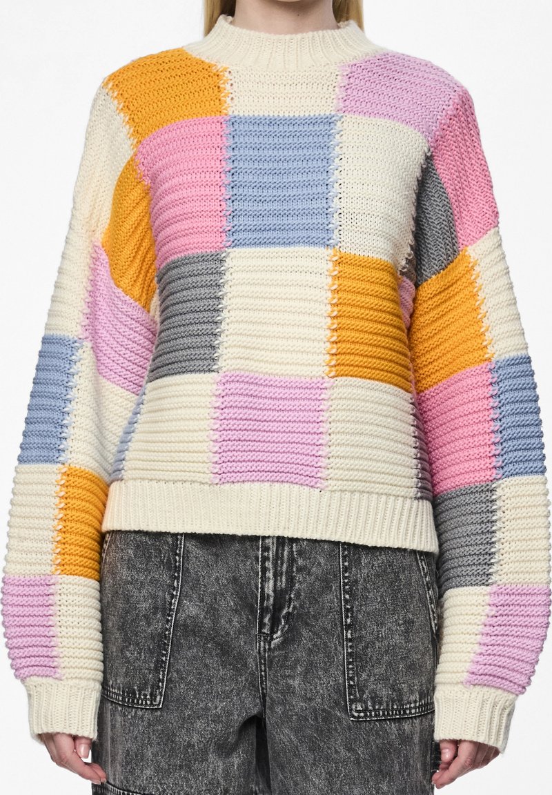 Pull en maille épaisse avec de grands carrés côtelés crème, rose, bleu, gris et orange, associé à un jean noir délavé à l'acide.