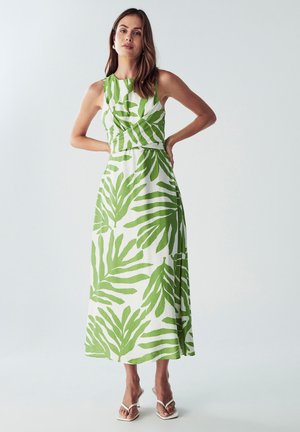 White maxi dress with large green leaf print, sleeveless design, fitted bodice, and flowing skirt. Paired with white heeled sandals.