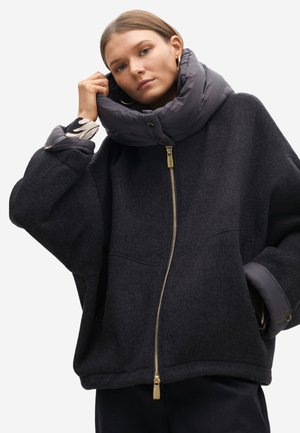 Chaqueta negra de gran tamaño con un cuello acolchado gris, cremallera dorada y grandes bolsillos laterales. La tela es una mezcla de lana y sintético.