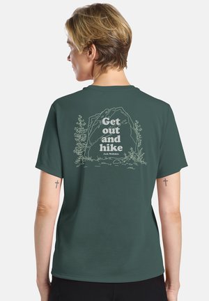 Person mit kurzen Haaren, die ein dunkelgrünes T-Shirt mit dem Text "Get out and hike" und einer Natur-Felsen-Illustration auf der Rückseite trägt.