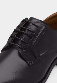 Μαύρο γυαλιστερό δερμάτινο παπούτσι με κλειστό κορδόνι, λείο επάνω μέρος και καφέ εσωτερική επένδυση, σε κοντινή λήψη σε λευκό φόντο.