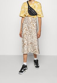 Gul grafisk t-shirt med vit text, svart midjeväska, midjelång krämfärgad kjol med bruna prickar, och svarta sportskor. Vita strumpor.