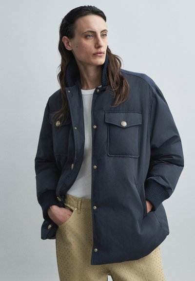 Navy blue quilted jacket with snap buttons, two front pockets, ribbed collar and cuffs, worn over a white top and patterned beige trousers.