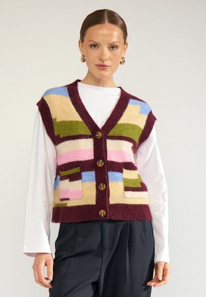 Femme portant un gilet en tricot multicolore boutonné avec des motifs géométriques par-dessus une chemise blanche à manches longues et un pantalon noir sur un fond uni.