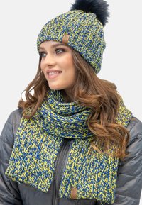 Strickmütze und Schal-Set in Blau und Gelb, mit Pompon, strukturiertem Muster und Leder-Logoschild. Getragen mit einer dunklen Jacke.