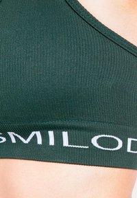 Skogsgrön ribbad sport-bh med ett brett vitt elastiskt band med varumärkeslogga längst ner. Designen har en elegant, minimalistisk form.