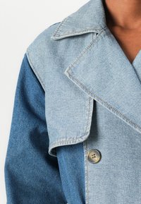 Blue denim jacket featuring a layered collar design, contrasting shades, a large lapel, and a brown button detail. Smooth texture.
