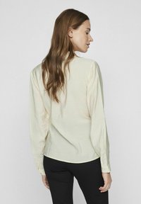 Blouse à manches longues, de couleur jaune clair, avec une coupe décontractée et une texture lisse, présente un ourlet droit et des poignets à bouton. Portée avec un pantalon noir.
