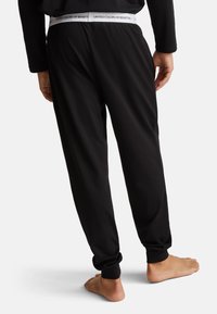 Zwarte joggingsbroek met elastische tailleband, voorzien van een witte logoband. De boorden zijn taps toelopend en de stof lijkt zacht en glad te zijn.
