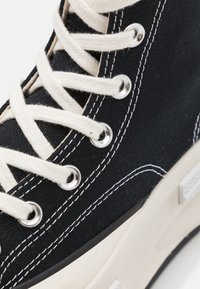 Zwarte canvas sneaker met witte veters, zilveren oogjes en contrasterende witte stiksels. Rubberen zool met een gestructureerd patroon.