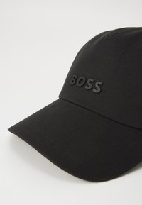 Svart cap laget av stoff med en strukturert design. Har en buet skygge og en hevet, svart "BOSS"-logo foran.
