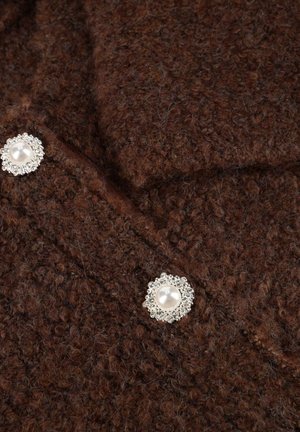 Bruine gestructureerde stof met duidelijke diepte; heeft twee decoratieve knopen met parelcentra omgeven door zilveren accenten.