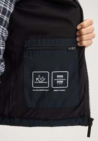 Czarna, wodoodporna i oddychająca podszewka kurtki z zamkniętą kieszenią, na której widnieją białe ikony symbolizujące wodoodporność i oddychalność.