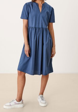 Kvinde iført knælang, blå kjole med korte ærmer og indsnævret talje, kombineret med hvide og lilla sneakers, stående på beige gulv.