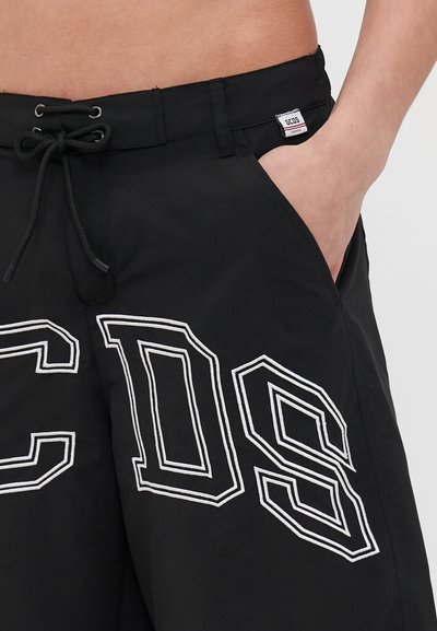 Černé kraťasy s páskem na stažení, bočními kapsami a velkým bílým vyšitým nápisem. Materiál se zdá být hladký s mírným leskem.