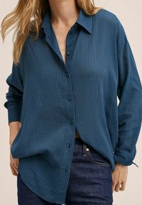 Kvindelig iført en løs, langærmet blå skjorte med lodrette striber og mørkeblå denimjeans, med hænderne delvist i lommerne.