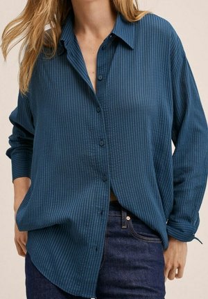 Kvindelig iført en løs, langærmet blå skjorte med lodrette striber og mørkeblå denimjeans, med hænderne delvist i lommerne.