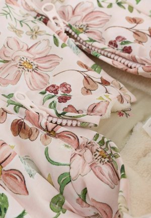 Látka s květinovým vzorem v jemné růžové barvě se zelenými akcenty; obsahuje zipy a nařasené rukávy, doplněná béžovou texturovanou podšívkou.