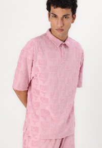 Pink short-sleeve polo shirt made of textured fabric, featuring a pattern of raised squares with a logo detail throughout. Buttoned collar.