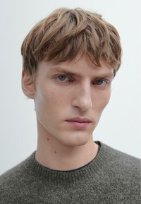 Primo piano di un modello maschile con capelli corti castano chiaro e occhi azzurri penetranti, che indossa un maglione verde scuro con trama e collo a coste.