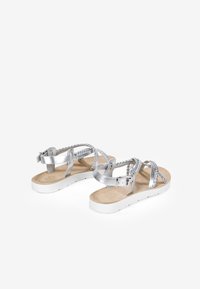 Sandalia plateada con correas trenzadas, hebilla ajustable y suela blanca plana. La plantilla suave presenta una textura en tono marrón claro.