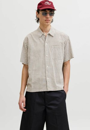 Junger Mann mit kurzärmeligem beigem kariertem Hemd, schwarzen Hosen, weinroter Mütze und rechteckigen dunklen Sonnenbrillen, stehend mit einer Hand in der Tasche.