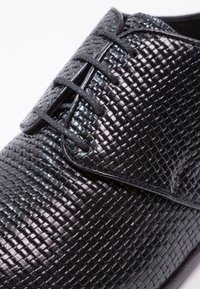 Chaussures en cuir tressé noir avec une finition brillante, dotées d'un design à lacets et d'une surface texturée. Les détails comprennent des bords lisses et des œillets.