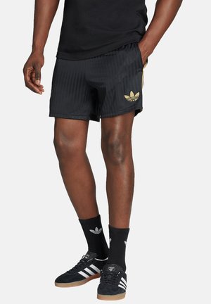 Shorts sportivi neri con strisce verticali, vita elastica e dettagli in logo dorato. Indossati con scarpe da ginnastica nere e calze con logo.