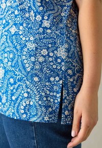 Blå blommönstrad topp med vita mönster. Har en sidoreva och en mjuk textur. Bärs med mörkblå denimjeans.