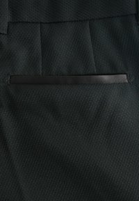 Tissu noir à finition texturée, avec une poche plate délimitée par un fin ruban noir, et des motifs diagonaux subtils.
