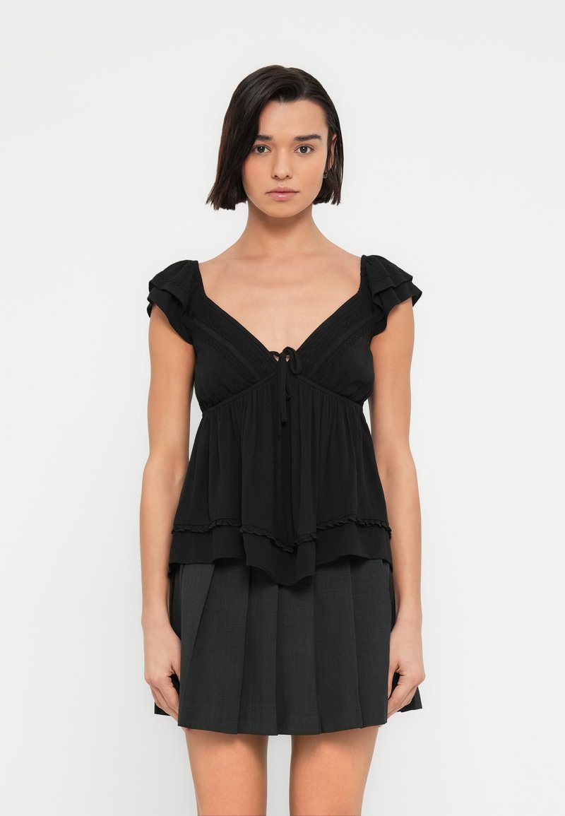 Μαύρο τοπ με κοντά βολάν στα μανίκια, V-λαιμόκοψη και λεπτομέρεια δέσιμου εμπρός, με στρωματοποιημένο ύφασμα και φραφλέ άκρα.