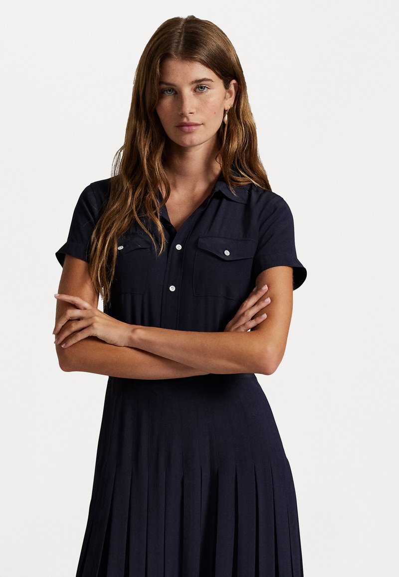Vrouw met lang bruin haar, gekleed in een marineblauw jurkje met korte mouwen en knopen, met borstzakken, staande met gekruiste armen tegen een witte achtergrond.