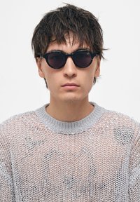 Mustat pyöreät aurinkolasit tummilla linsseillä, hopeiset metalliosat ja tekstuuriharmaa neulepusero leveällä pääntiellä.