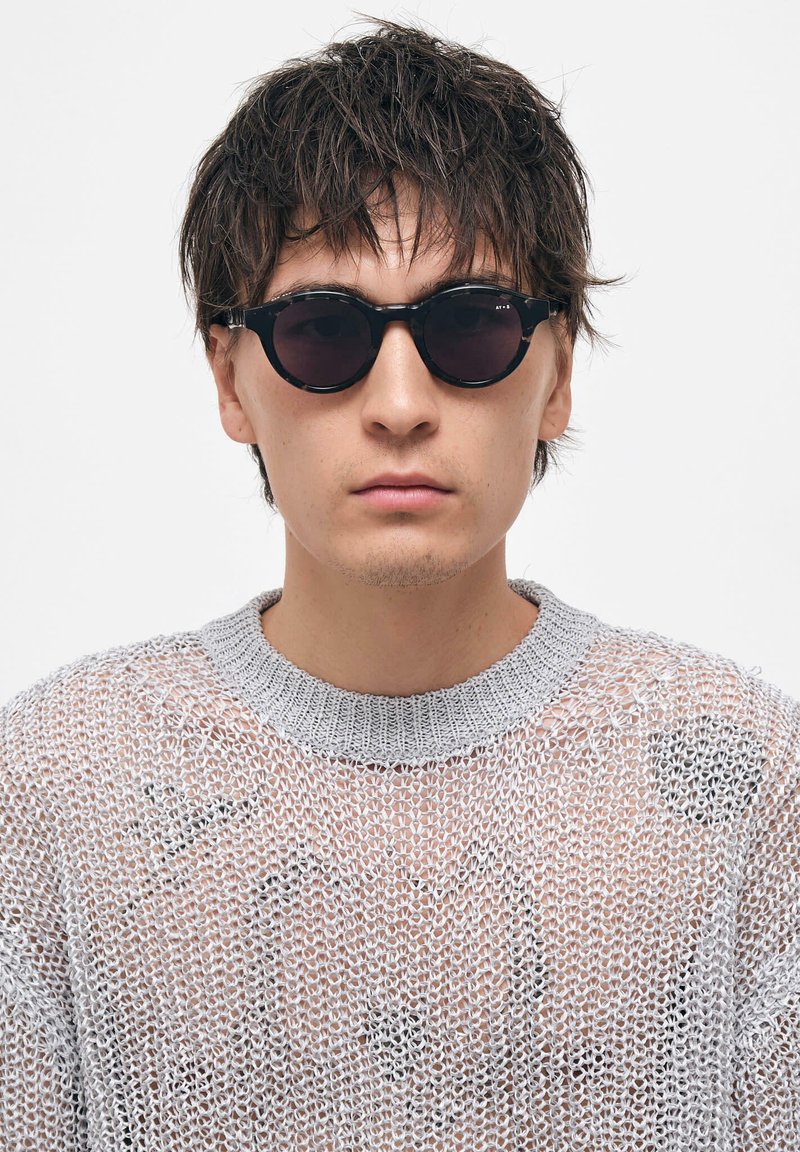 Mustat pyöreät aurinkolasit tummilla linsseillä, hopeiset metalliosat ja tekstuuriharmaa neulepusero leveällä pääntiellä.