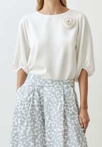 Weißes Blusenshirt mit kurzen, puffigen Ärmeln und einer Stoffrose als Akzent. Kombiniert mit einem hellblauen Rock mit gepunktetem Muster und Falten.