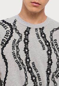 Graues Baumwoll-T-Shirt mit schwarzen Ketten- und Textmustern, Rundhalsausschnitt und kurzen Ärmeln. Der Text beinhaltet "Versace Jeans Couture."