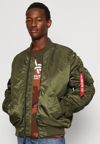 Olive green bomber jacket with a zip front, ribbed cuffs, and a pocket on the sleeve featuring a red pull tag. Worn over a brown camo shirt.