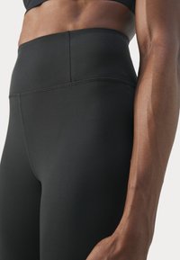 Magas derekú fekete leggings rugalmas anyagból, sima felülettel és lapos varrásokkal, a derék és a kar közeli képen látható.