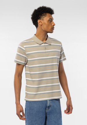 Ung mann med kort krøllete hår iført en beige, hvit og blåstripet poloskjorte og blå jeans, ser mot høyre.
