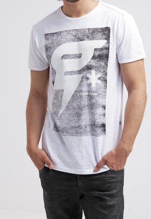 Homme portant un T-shirt blanc à manches courtes avec un grand « F » stylisé et un motif étoile, et un pantalon noir, les mains dans les poches.