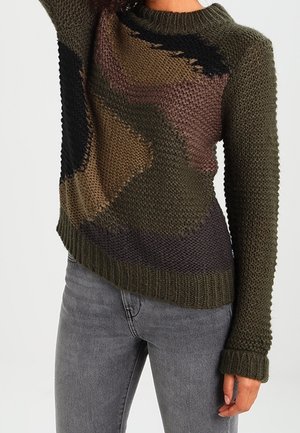 Žena v dlouhém pleteném svetru s texturovaným, vícebarevným geometrickým vzorem v zelené, hnědé a černé barvě, doplněném šedými džínami.