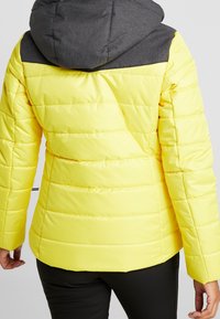 Gul pufferjacka med grå huva, med quiltad textur, dragkedjestängning och sömlösa designelement på axlarna och ärmarna.